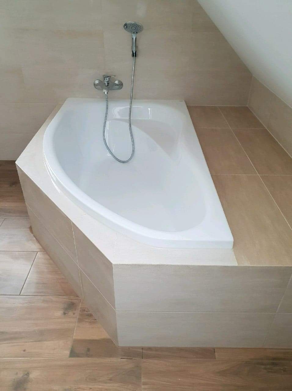 Biała narożna wanna obudowana jasnymi płytkami ceramicznymi w łazience na poddaszu, widoczna bateria i słuchawka prysznicowa.