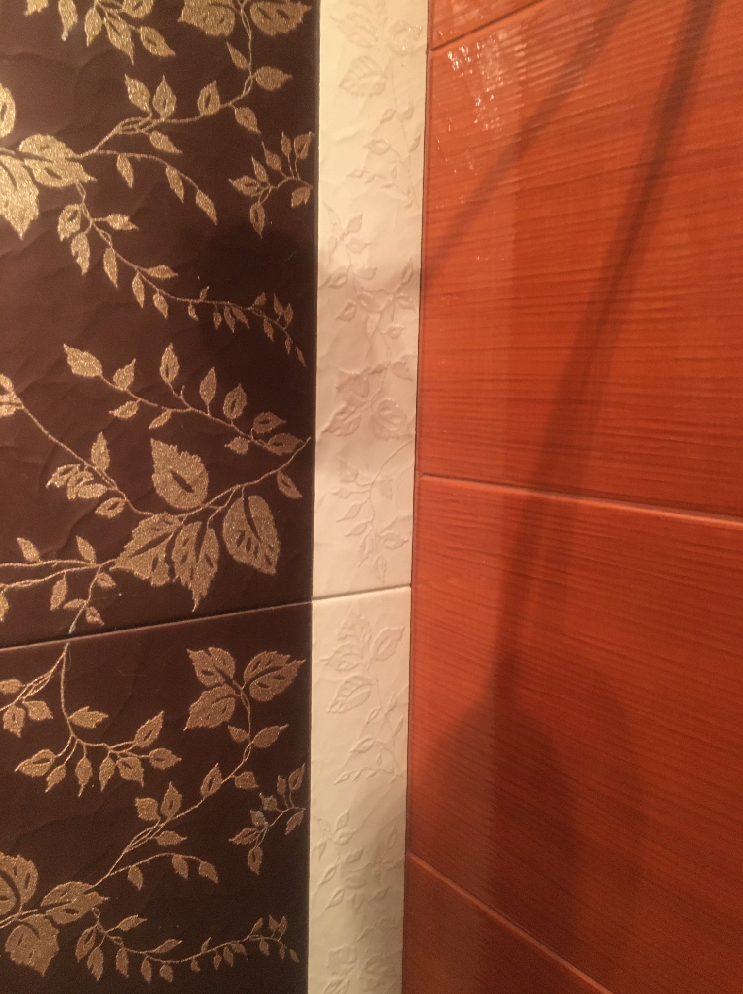 Detal płytek ściennych: brązowe z motywem złotych liści, białe z delikatnym wzorem roślinnym i pomarańczowe z poziomymi prążkami imitującymi drewno.