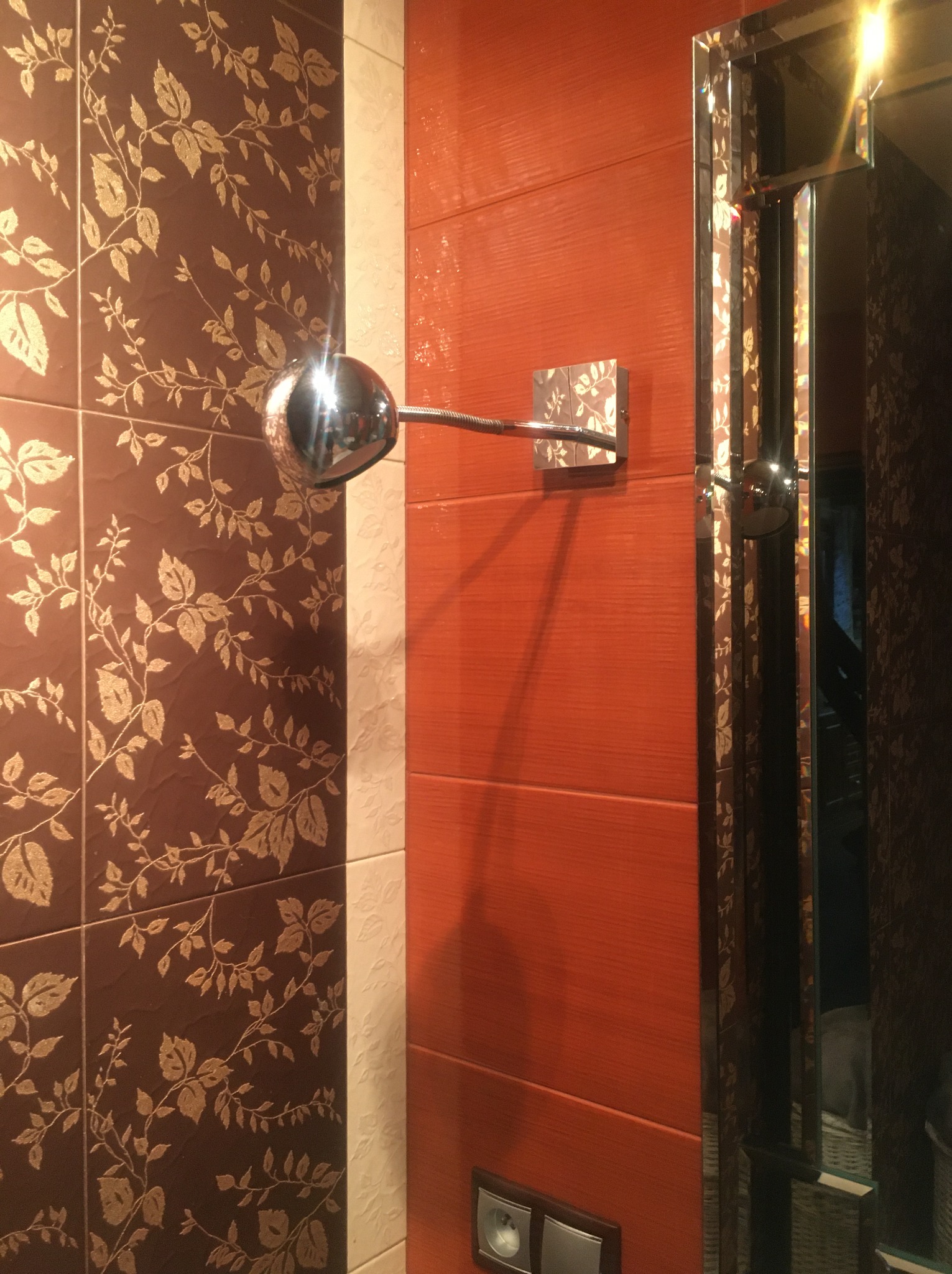 Detal ściany łazienki z brązowymi i pomarańczowymi płytkami z motywem roślinnym, chromowana lampa ścienna i lustro z fazowanymi krawędziami.