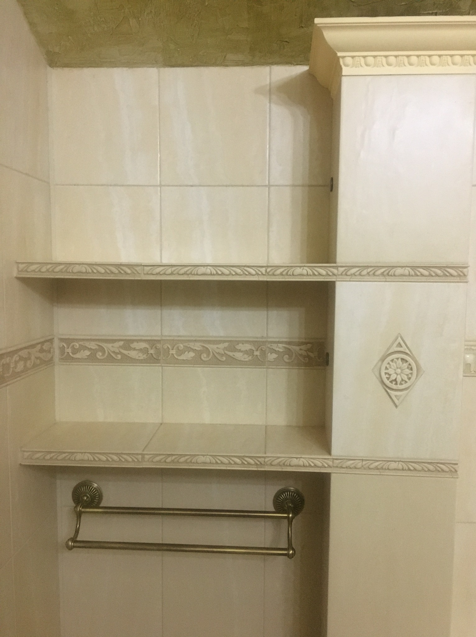 Wykończona łazienka z beżowymi płytkami, dwoma półkami z dekoracyjnym obramowaniem, szafką z ornamentem i mosiężnym wieszakiem na ręczniki.