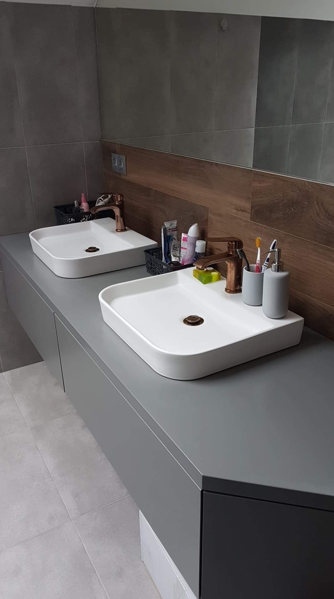 Dwie białe, kwadratowe umywalki nablatowe z miedzianą armaturą na szarym blacie w nowoczesnej łazience z szarymi płytkami i drewnopodobną wstawką.