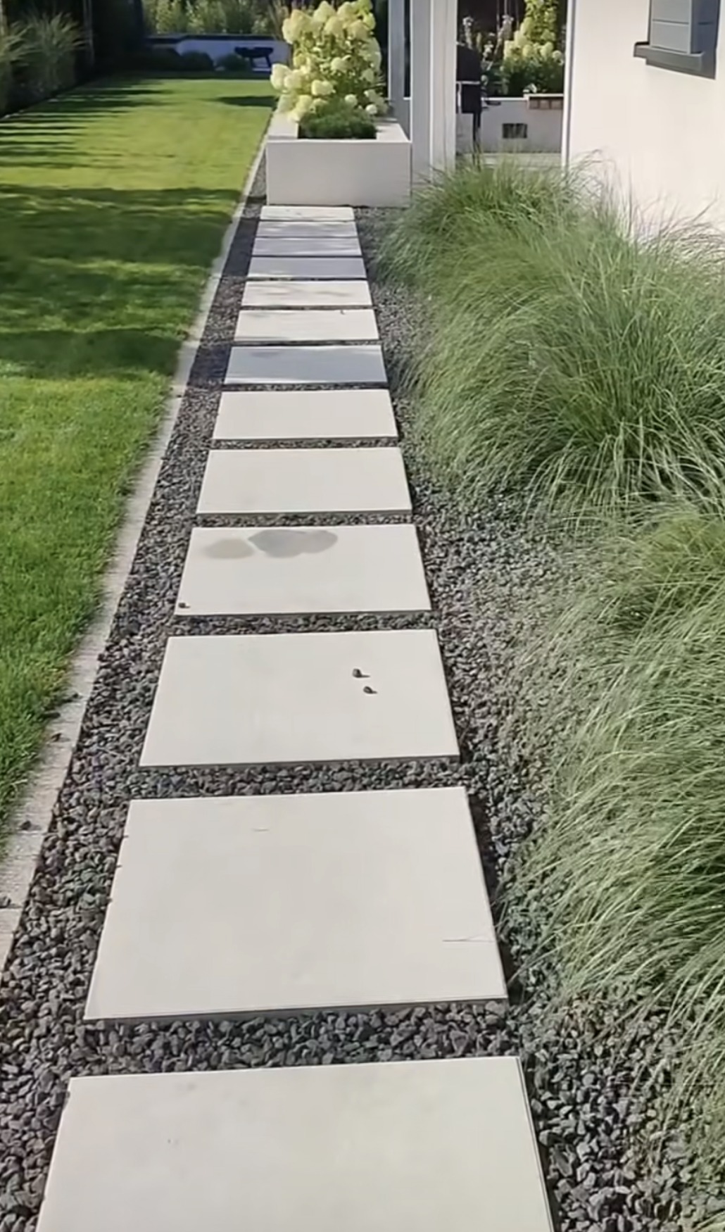 Ścieżka z jasnych płyt w ogrodzie, otoczona szarym żwirem i ozdobną trawą, prowadząca w stronę domu z białymi elementami architektonicznymi.