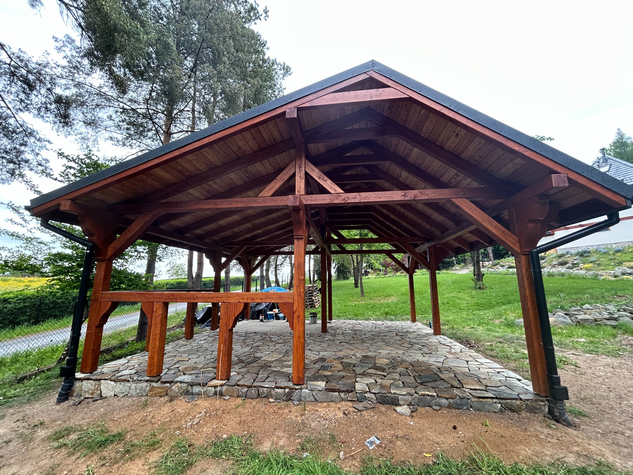 Drewniana altana z kamienną podłogą na zewnątrz, widok z boku. Konstrukcja z ciemnego drewna, kamienna posadzka z nieregularnych płyt, zielone otoczenie.