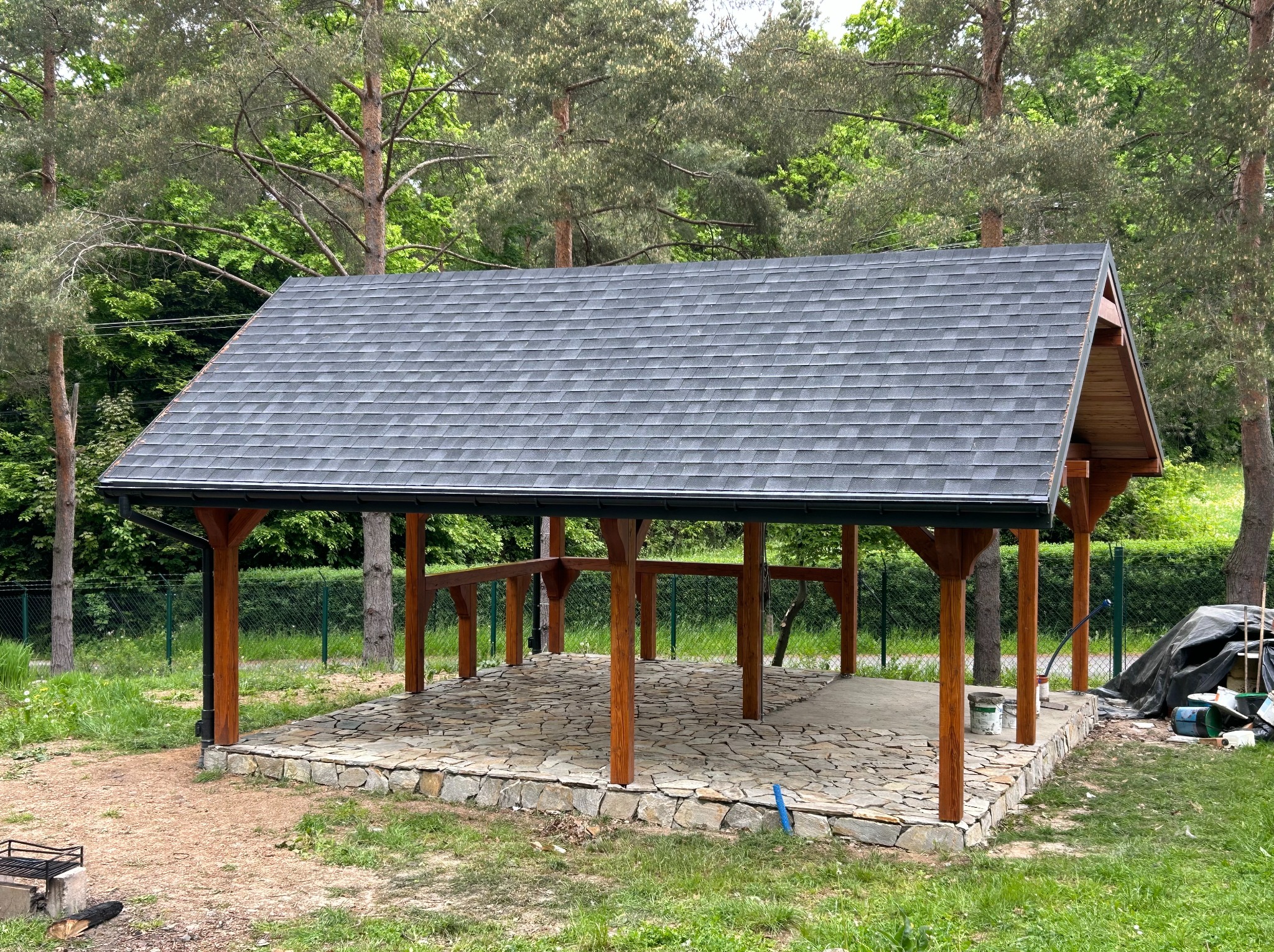 Zadaszona wiata ogrodowa z drewnianymi słupami i kamienną podłogą, otoczona zielenią. Widoczne puszki z farbą i narzędzia, sugerujące prace wykończeniowe.