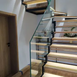 Nowoczesne schody z drewnianymi stopniami, czarną metalową konstrukcją i szklaną balustradą, podświetlane od spodu, z dekoracjami na półkach i kartonami zabezpieczającymi na podłodze.