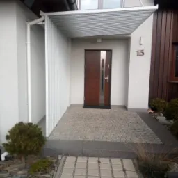 Nowoczesne wejście do domu z zadaszeniem z białych, poziomych listew, brązowe drzwi z metalowymi akcentami, numer domu '15' na białej ścianie, kamienna nawierzchnia i kostka brukowa.