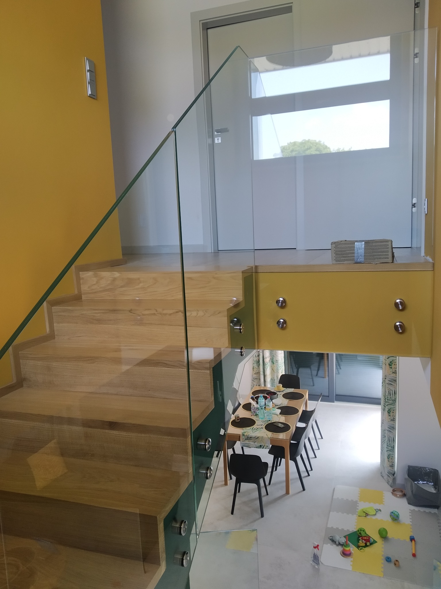 Wnętrze domu z drewnianymi schodami zabezpieczonymi szklaną balustradą, mocowaną za pomocą metalowych uchwytów. W tle widoczny stół jadalny i pokój dziecięcy z matą edukacyjną.