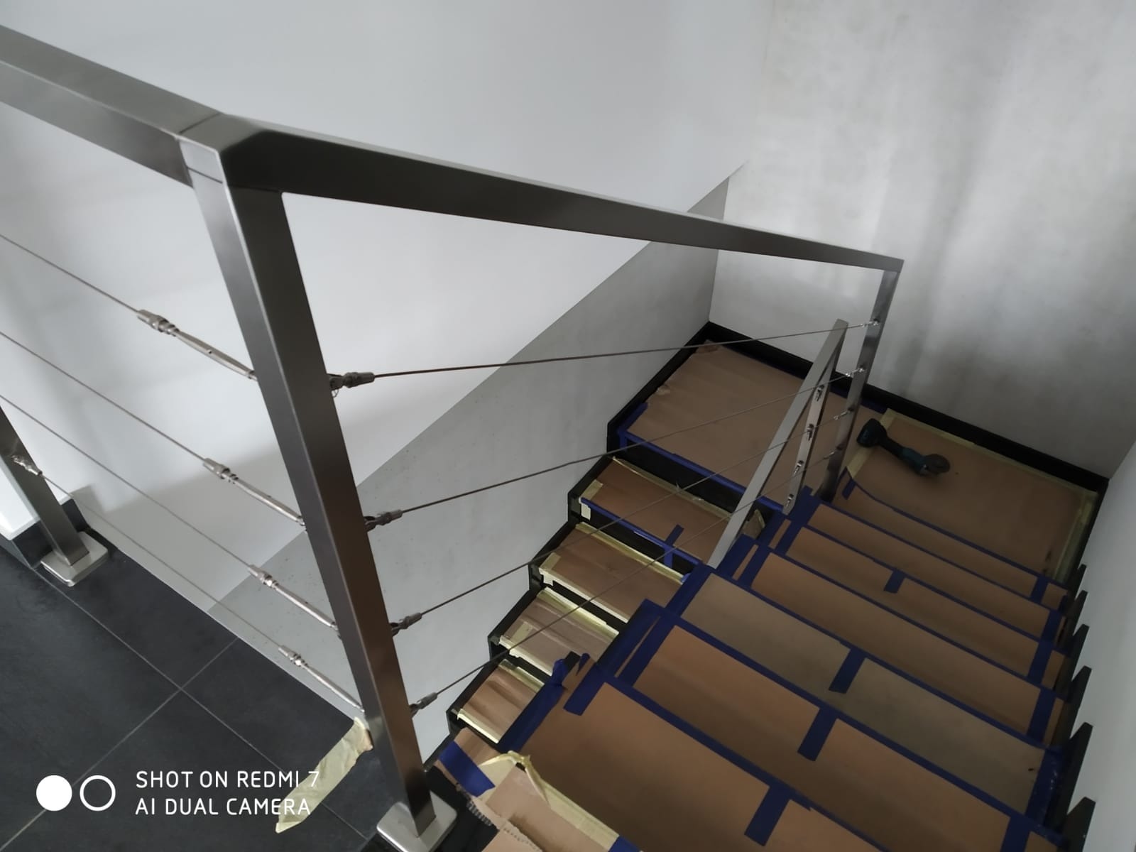 Metalowa poręcz schodowa z linkami stalowymi, widok z góry na schody zabezpieczone kartonem i taśmą malarską, w tle biała ściana.