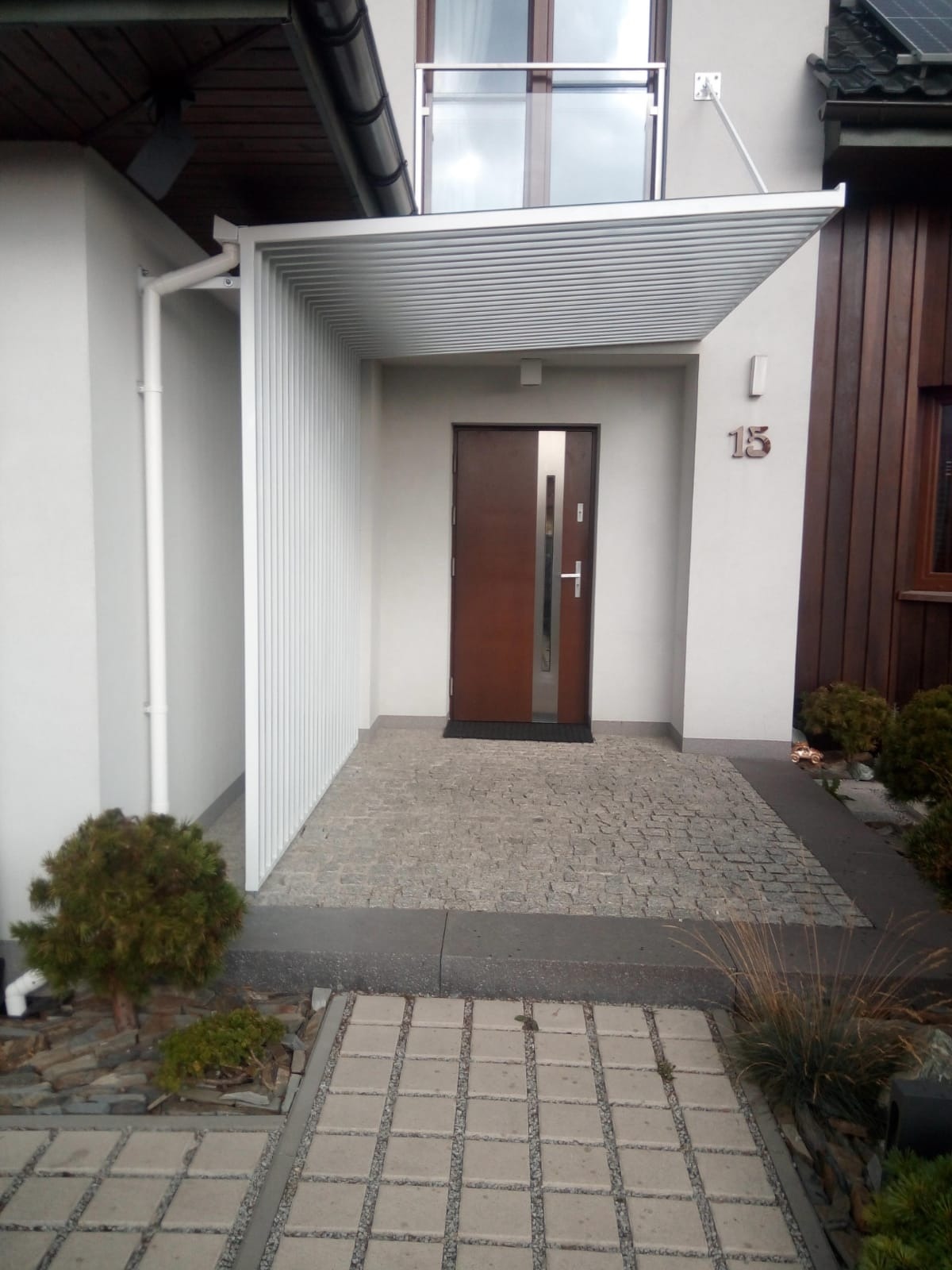 Nowoczesne wejście do domu z zadaszeniem z białych, poziomych listew, brązowe drzwi z metalowymi akcentami, numer domu '15' na białej ścianie, kamienna nawierzchnia i kostka brukowa.