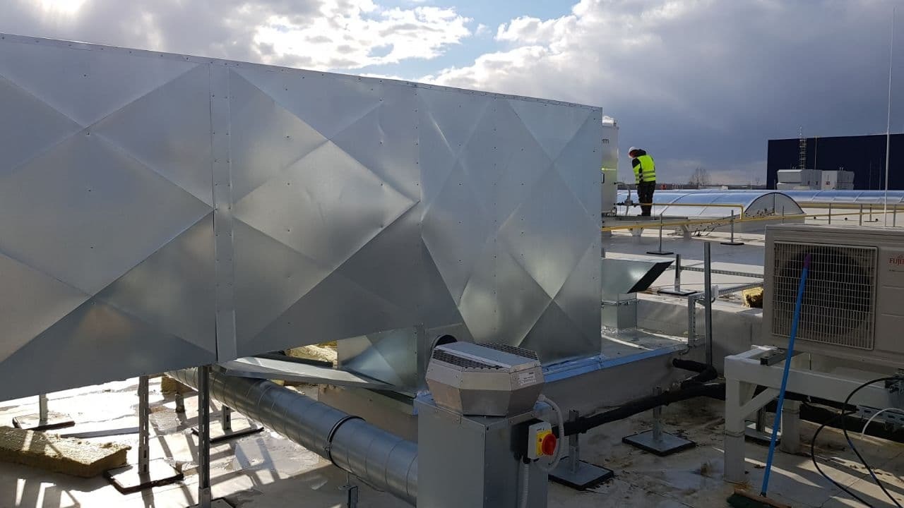 Instalacja wentylacyjna na dachu budynku komercyjnego z widocznym pracownikiem w kamizelce odblaskowej i klimatyzatorem Fujitsu w tle.