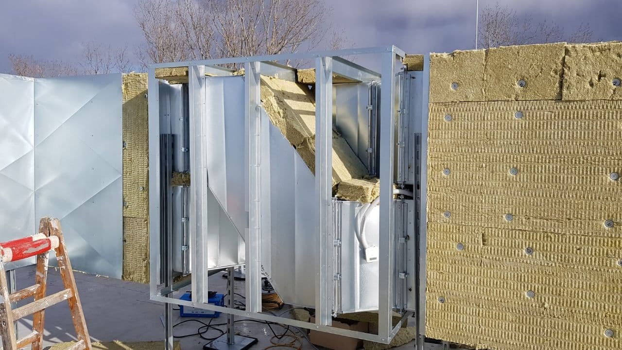 Widok z zewnątrz na fragment systemu kanałów wentylacyjnych w trakcie montażu, z widoczną izolacją termiczną i konstrukcją wsporczą, drabina malarska w lewym dolnym rogu.