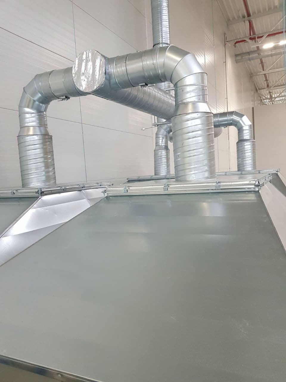 System kanałów wentylacyjnych z blachy ocynkowanej, widok z dołu na elementy łączeniowe i izolację na jednym z odcinków, w tle hala przemysłowa.