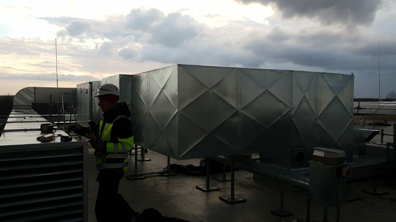 Pracownik w kasku montuje system kanałów wentylacyjnych na dachu budynku komercyjnego; widoczne elementy systemu o strukturze romboidalnej i narzędzia pracy.