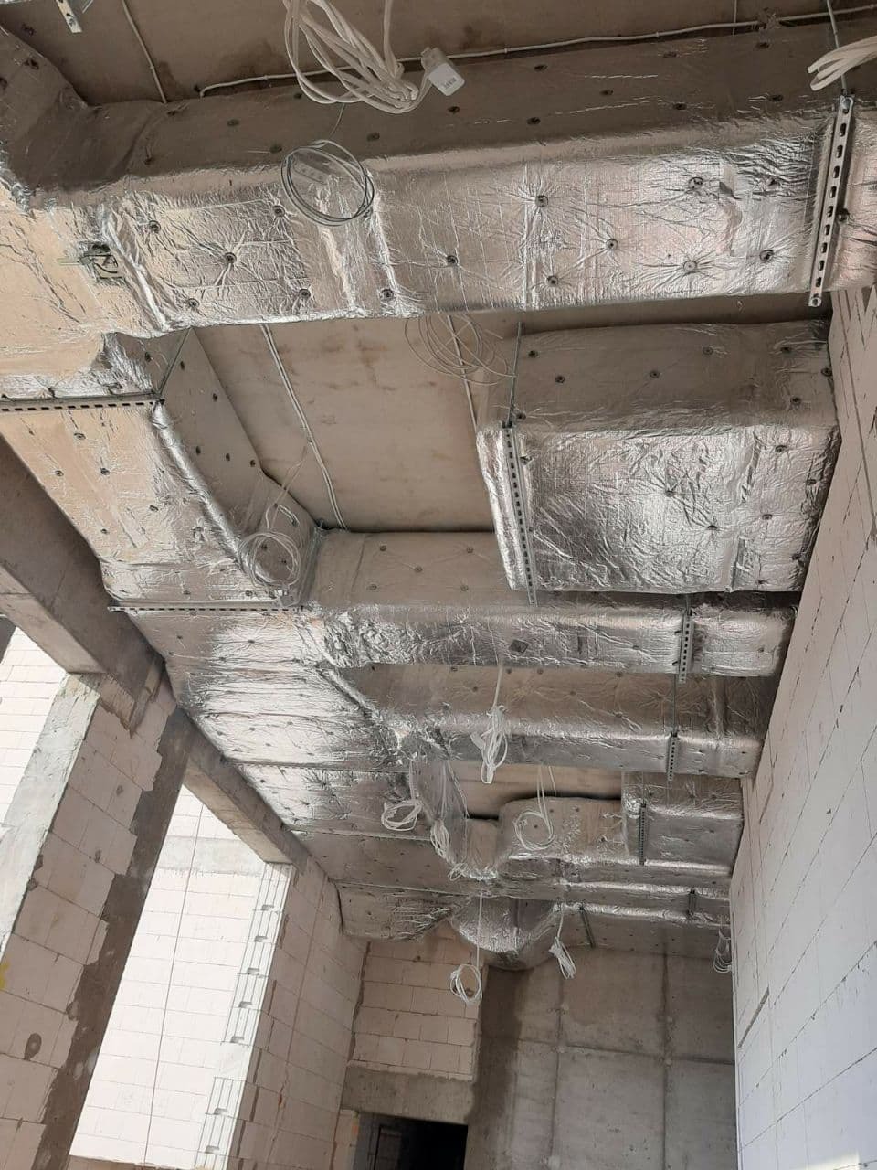 Montaż izolowanych kanałów wentylacyjnych pod sufitem w surowym stanie budynku, widoczne przewody elektryczne i elementy konstrukcyjne.