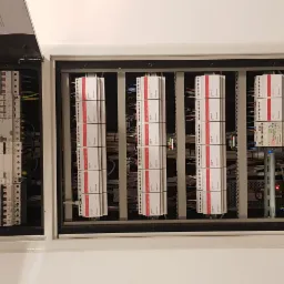 Otwarta szafa rozdzielcza z widocznymi bezpiecznikami, modułami sterującymi i okablowaniem w kolorach niebieskim, żółtym i czerwonym, zamontowana na białej ścianie.