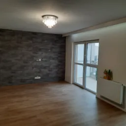 Pusty pokój po remoncie z drewnianą podłogą, szarą ścianą z imitacją kamienia, białymi ścianami, grzejnikiem i ozdobnym żyrandolem.