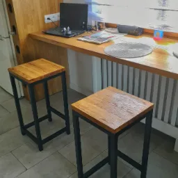 Dwa nowoczesne stołki barowe z drewnianymi siedziskami i czarnymi, metalowymi nogami, ustawione na jasnej, kafelkowej podłodze w Pyskowicach. W tle blat roboczy z laptopem i akcesoriami.