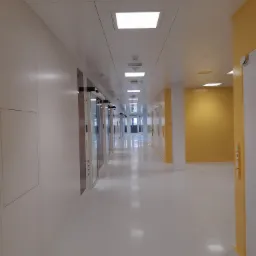 Długi korytarz z panelami ściennymi i sufitem podwieszanym z wbudowanym oświetleniem, widoczne drzwi przesuwne i żółte akcenty kolorystyczne na ścianach i ościeżnicach.