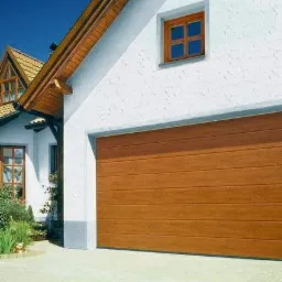 Zamknięta, segmentowa brama garażowa w kolorze jasnego drewna, zamontowana w białym garażu z małym okienkiem. Widoczny fragment domu z brązowym dachem i oknami.