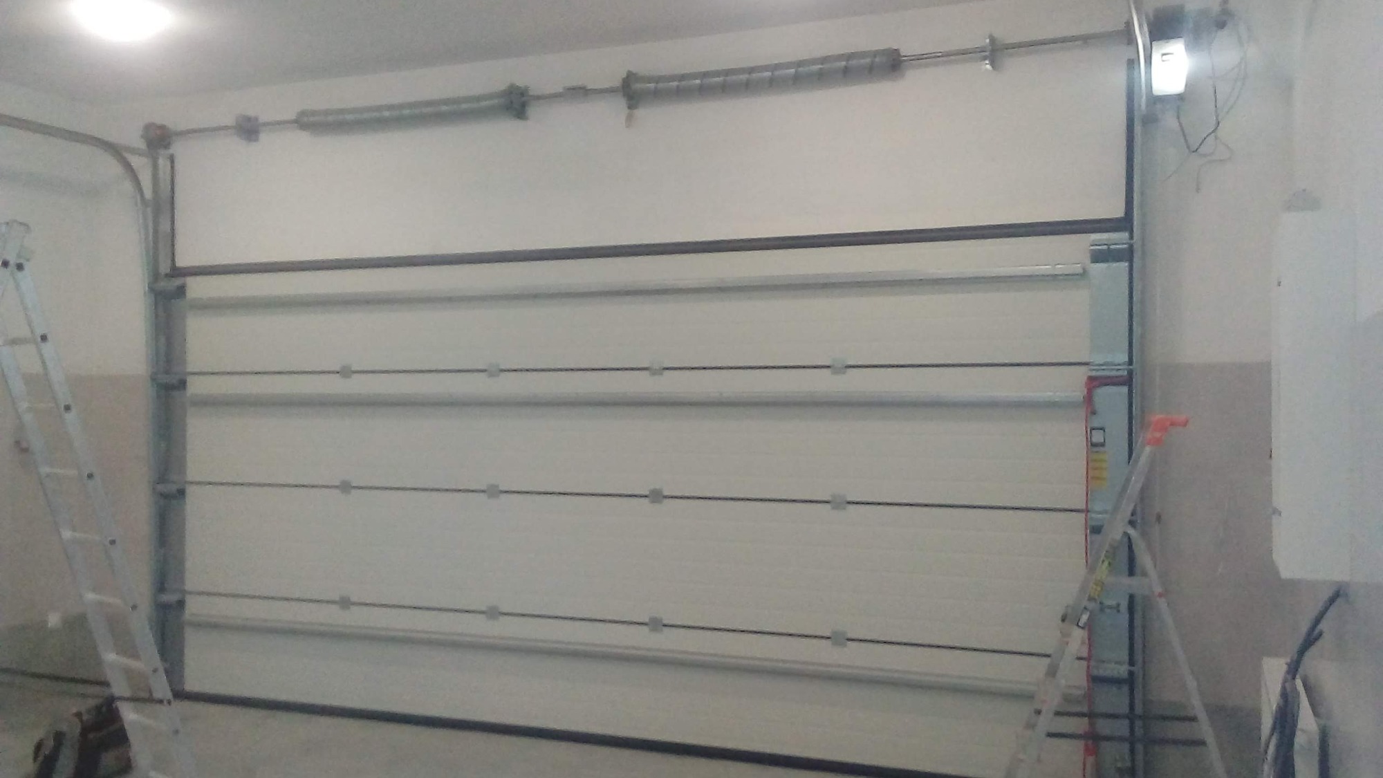 Montaż segmentowej bramy garażowej Ryterna w jasnym garażu, widoczne drabiny monterskie i sprężyny nad bramą.