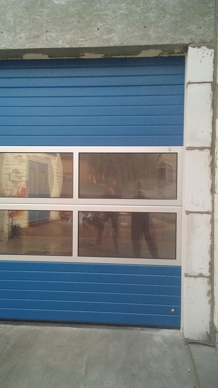 Niebieska segmentowa brama z czterema prostokątnymi oknami, w których odbija się otoczenie, widoczna betonowa rama budynku.