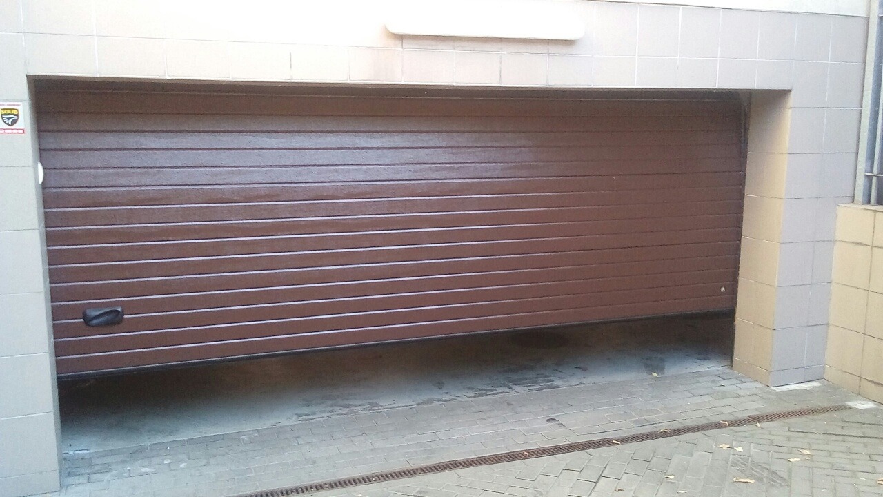 Częściowo otwarta, segmentowa brama garażowa Ryterna w kolorze brązowym, zamontowana w garażu w Sochaczewie, widoczny uchwyt bramy, szara kostka brukowa przed wjazdem.