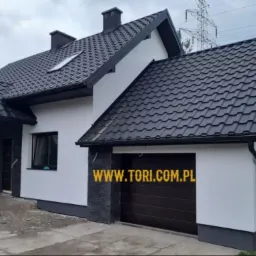 Nowoczesny dom jednorodzinny z ciemną, metalową dachówką, białymi ścianami z elementami kamiennymi i ciemną stolarką okienną oraz wstawioną bramą garażową.