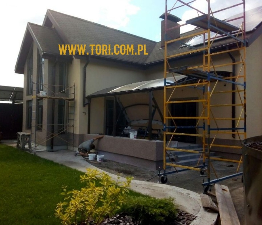 Dwupiętrowy dom z beżową elewacją w trakcie prac renowacyjnych, widoczne rusztowania i pracownik przy niskiej ścianie, zielony trawnik z przodu.
