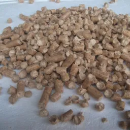 Kupa jasnobrązowych, cylindrycznych granulek paliwa na białym podłożu, widoczne nierówności i drobne uszkodzenia struktury.