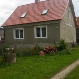 Dom jednorodzinny z nowym, czerwonym dachem z dachówki, widoczne dwa okna dachowe i komin. Na trawniku leżą stare dachówki. Dom otynkowany na szaro, z numerem 13 na rogu. Na pierwszym planie studnia.