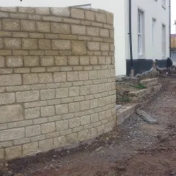 Zakrzywiona ściana z piaskowca z widocznymi spoinami, w tle budynek mieszkalny i plac budowy.