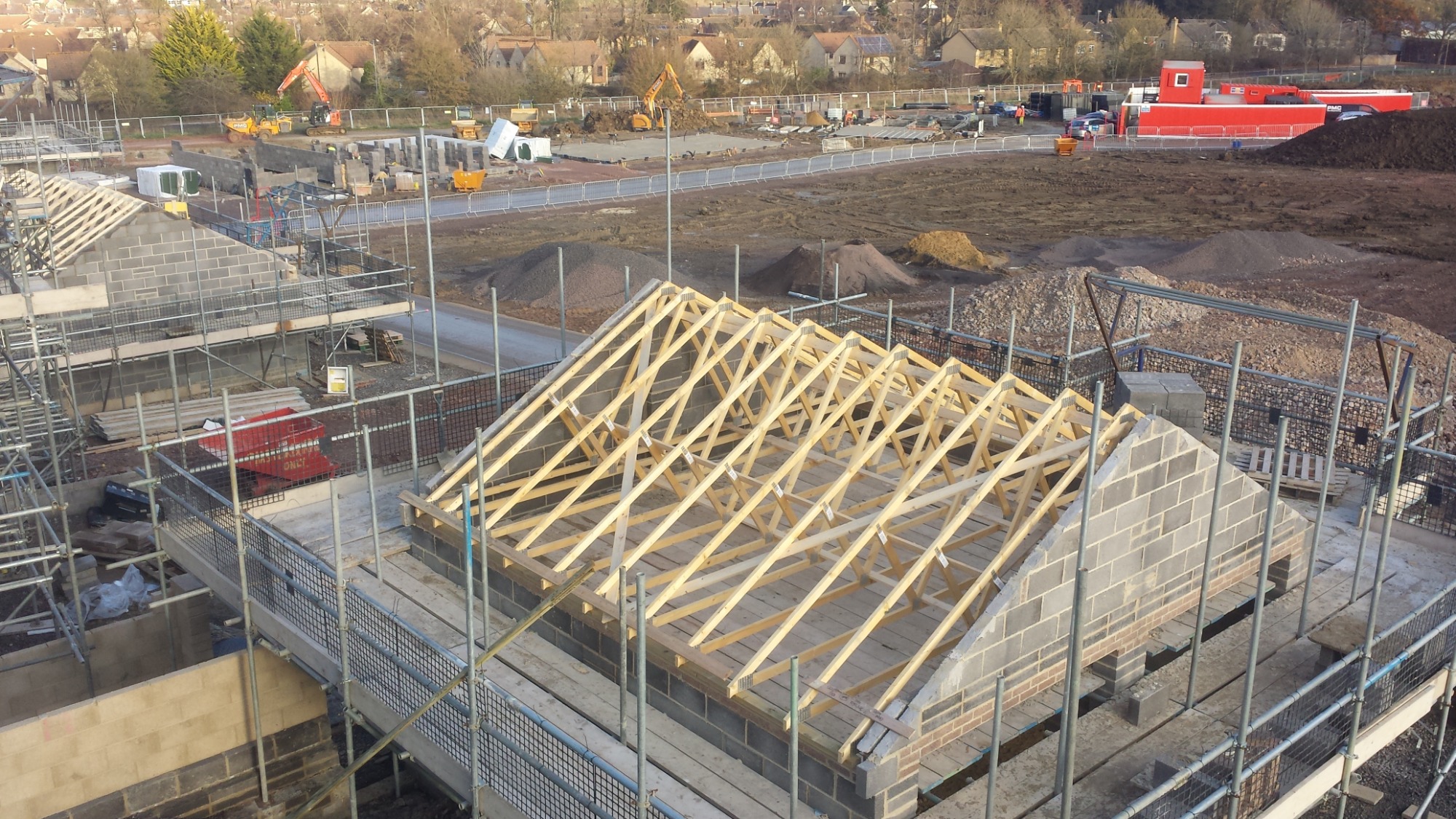 Wznoszenie konstrukcji dachu z drewnianych krokwi na murowanym budynku z bloczków, widoczne rusztowania i plac budowy z maszynami w tle.