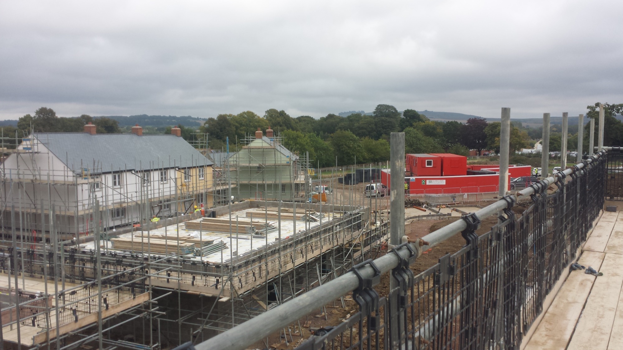 Budowa osiedla domów jednorodzinnych w trakcie realizacji, widoczne rusztowania, fundamenty, ściany wznoszone z bloczków, kontenery budowlane i teren przygotowany pod dalsze prace.