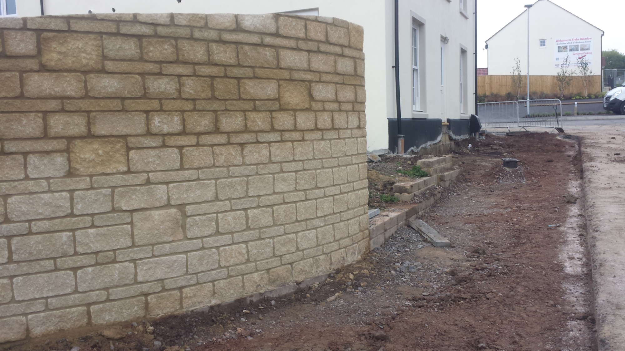 Zakrzywiona ściana z piaskowca z widocznymi spoinami, w tle budynek mieszkalny i plac budowy.