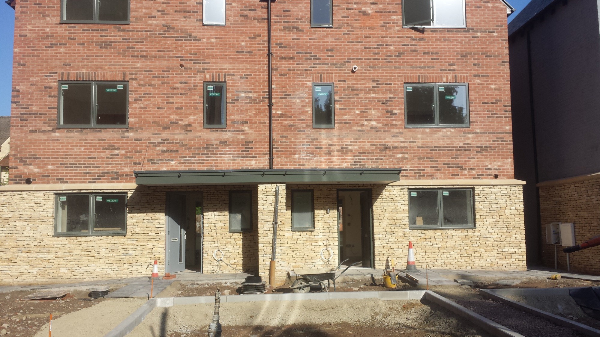 Dwukondygnacyjny budynek z cegły i kamienia, z szarymi oknami, w trakcie budowy lub remontu, z widocznymi materiałami budowlanymi i narzędziami na placu budowy.