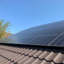 Panele słoneczne zamontowane na brązowym dachu z blachodachówki, widok z dołu, błękitne niebo w tle.