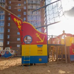 Kolorowy plac zabaw w kształcie statku pirackiego z żółtą kotwicą na tle budynku mieszkalnego.