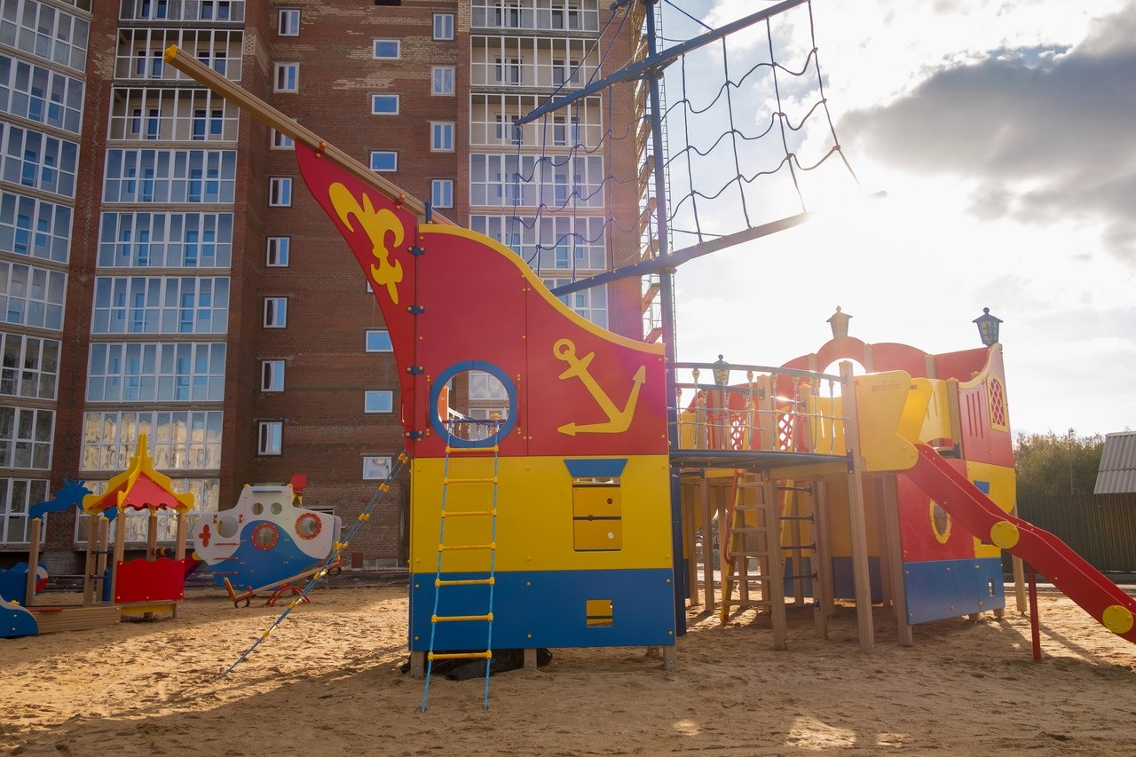 Kolorowy plac zabaw w kształcie statku pirackiego z żółtą kotwicą na tle budynku mieszkalnego.