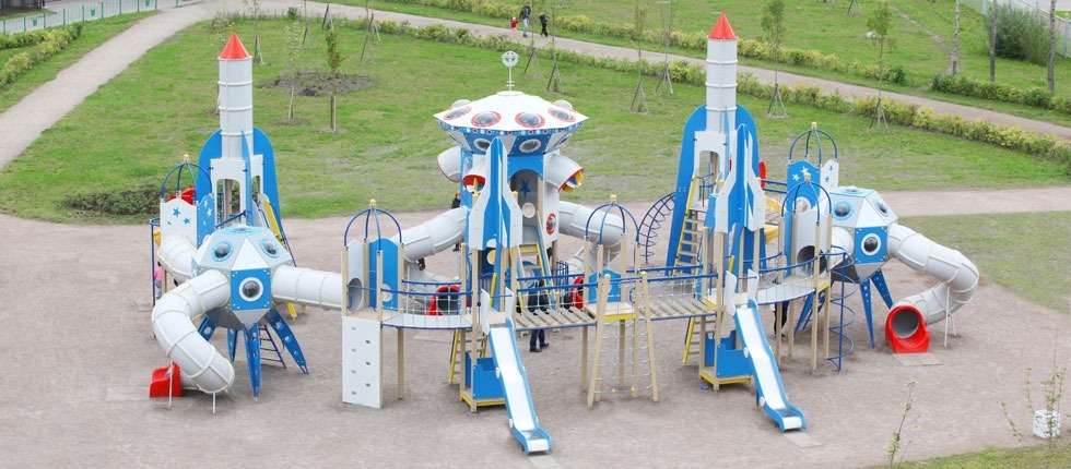 Kompleksowy plac zabaw w kształcie statków kosmicznych i rakiet z elementami w kolorze białym, niebieskim i czerwonym, zjeżdżalniami, drabinkami i tunelami, na piaszczystym podłożu i z zielenią w tle.