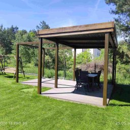 NA TARASIE Sp. z o.o. - Nowoczesna, ciemnobrązowa pergola z drewnianą podłogą i kompletem mebli ogrodowych na zadbanym trawniku, w tle zieleń drzew i plac zabaw.