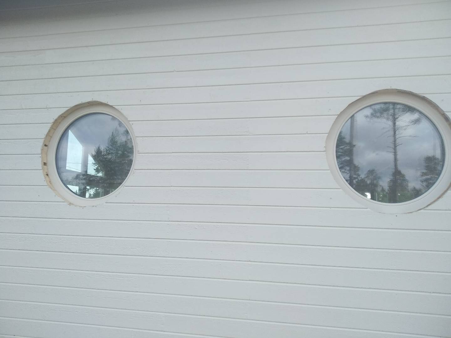 Fragment białej ściany z desek z dwoma okrągłymi oknami, w których odbijają się drzewa i niebo.
