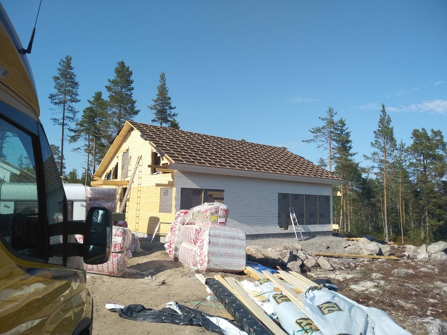 Budowa domu w technologii szkieletowej w otoczeniu drzew iglastych, widoczne materiały izolacyjne i elementy konstrukcyjne dachu, żółty bus w lewym dolnym rogu kadru.