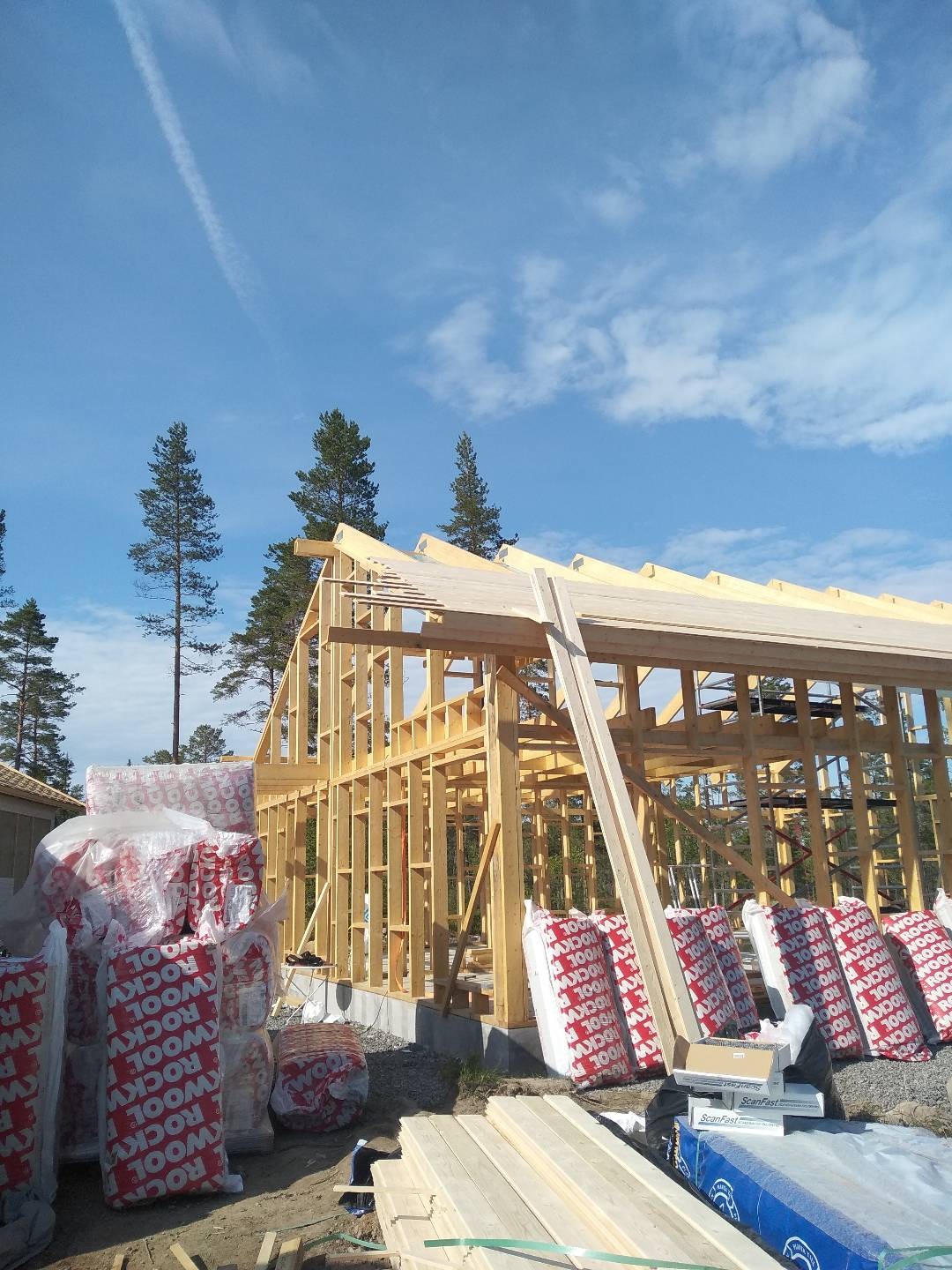 Wznoszenie drewnianej konstrukcji domu w technologii szkieletowej, widoczne belki, izolacja termiczna ROCKWOOL w opakowaniach i przygotowane deski, błękitne niebo w tle.