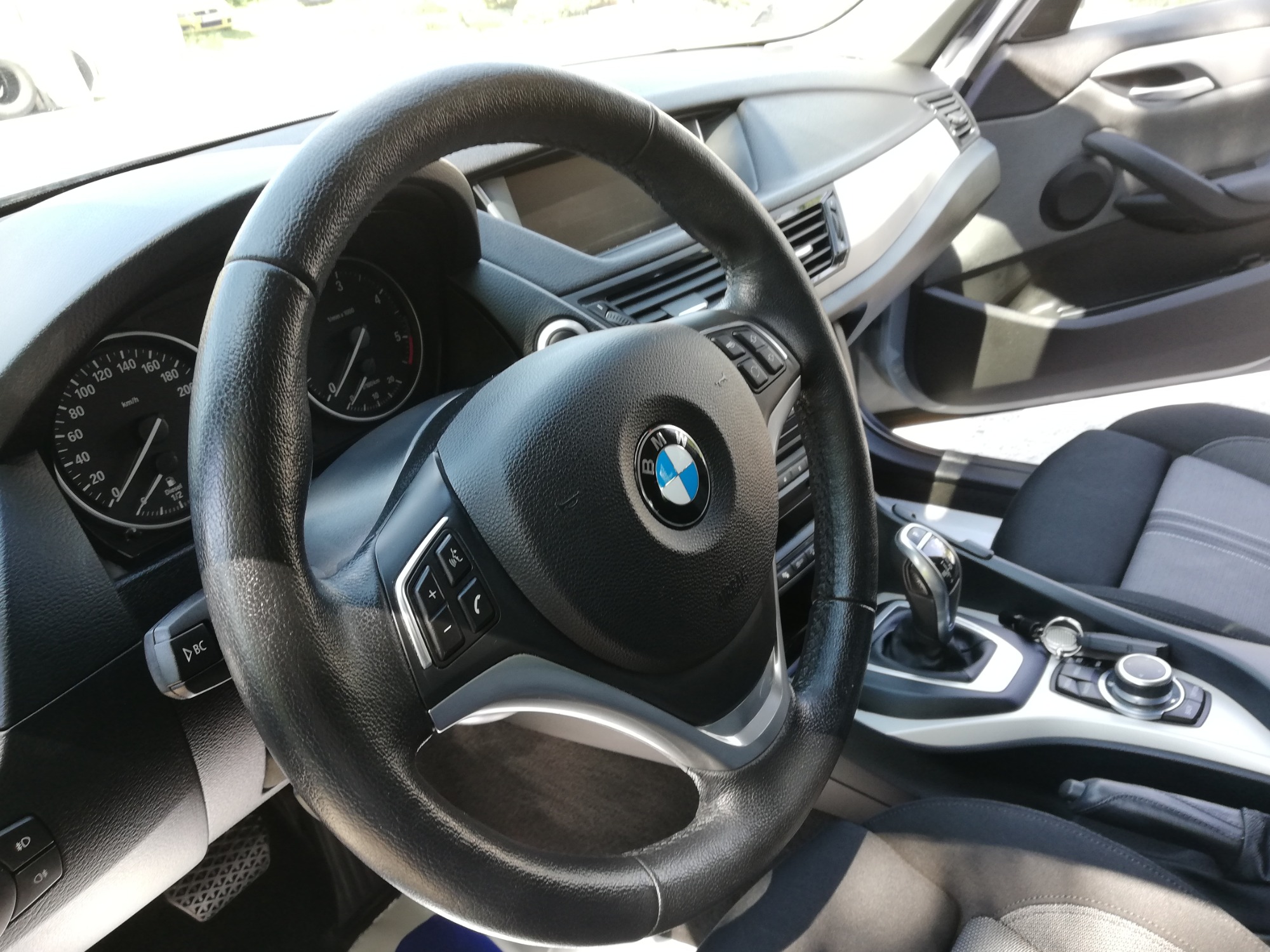 Wnętrze samochodu BMW, widok na kierownicę, deskę rozdzielczą i fotel kierowcy. Skórzana kierownica z logo BMW w centralnej części, panel sterowania radiem i tempomatem po lewej stronie. Widoczne...