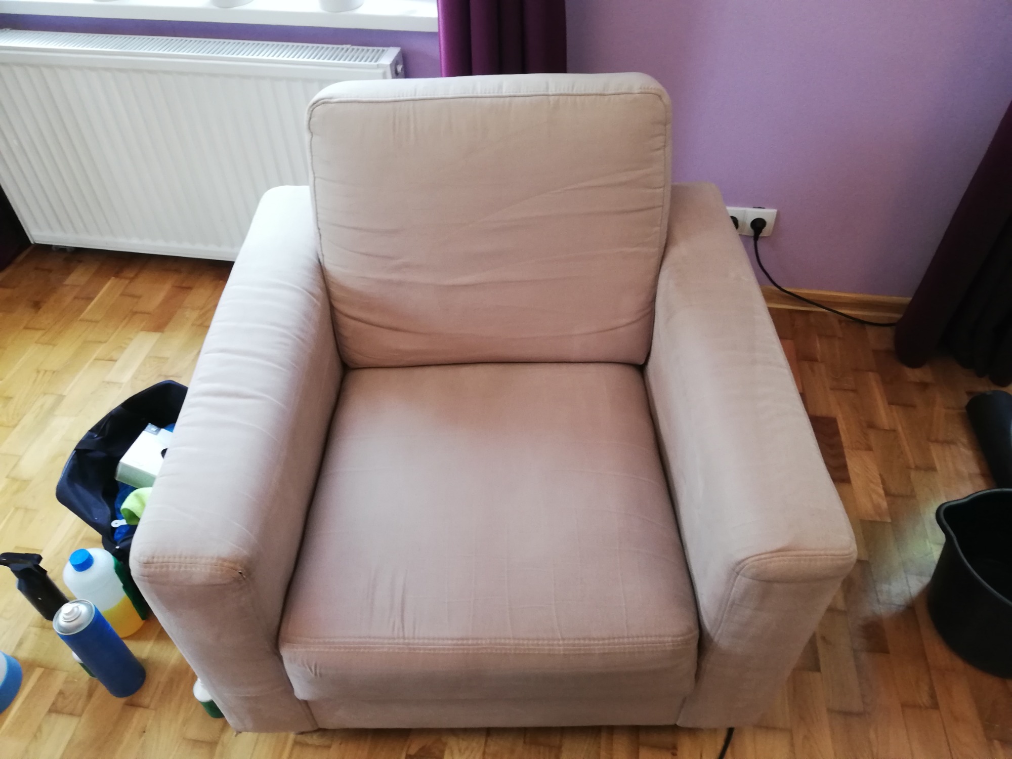 Jasny fotel z tapicerką po czyszczeniu, widoczne akcesoria do sprzątania obok, na tle drewnianej podłogi i fioletowej ściany.