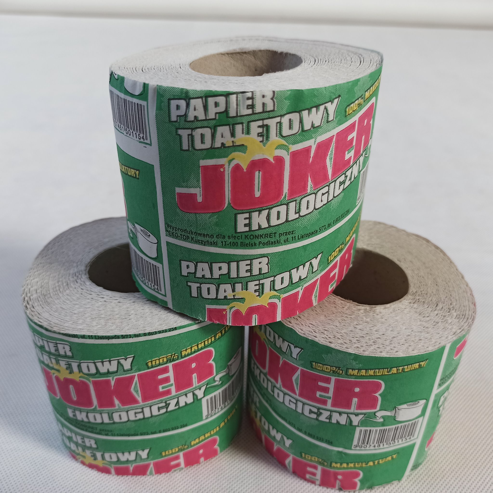 Trzy rolki makulaturowego papieru toaletowego marki Joker Ekologiczny, ułożone jedna na drugiej, z widocznym zielono-czerwonym opakowaniem i kodem kreskowym.