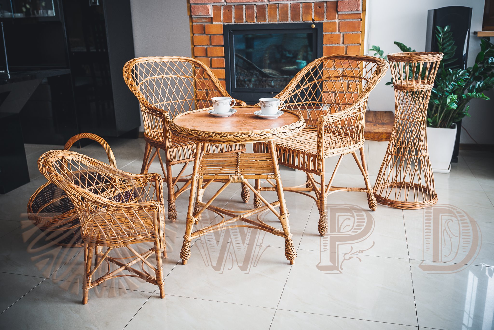 Wnętrze z wiklinowymi meblami: stolik z filiżankami, krzesła, kwietnik, kosz. W tle kominek z cegły i czarne meble.