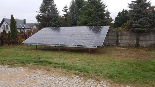 Instalacja paneli słonecznych na trawniku przed domem jednorodzinnym, widoczne drzewa i szary, pochmurny dzień.