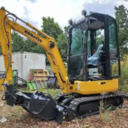 Żółta mini koparka Komatsu PC16R na ziemi z liśćmi, w tle biały kontener i drzewa.