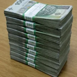 Stosy spiętych banknotów 100 złotych polskich na drewnianym blacie, widoczne banderole z białymi polami do opisu.